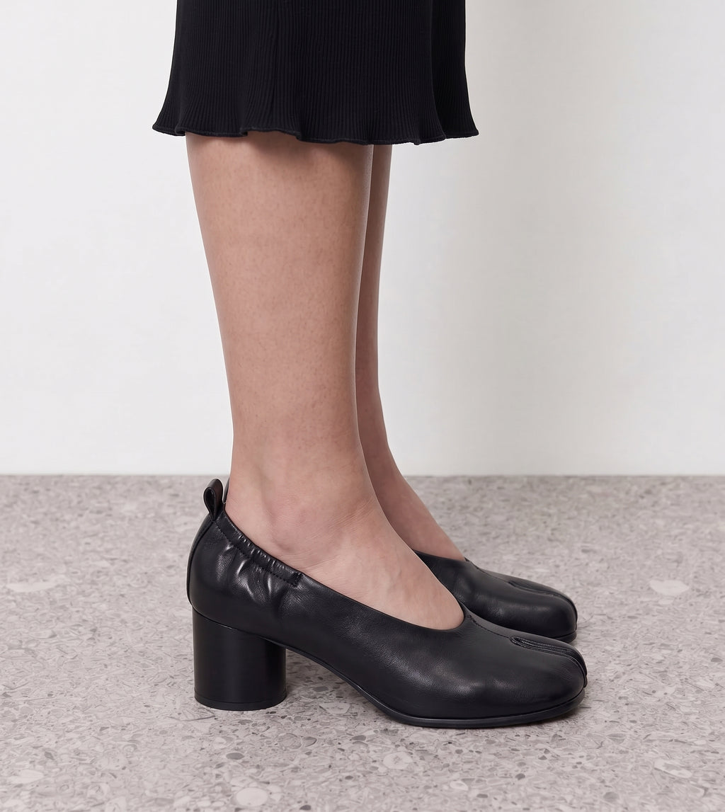 Side profile of a black tabi pump on a round block heel worn with a black skirt