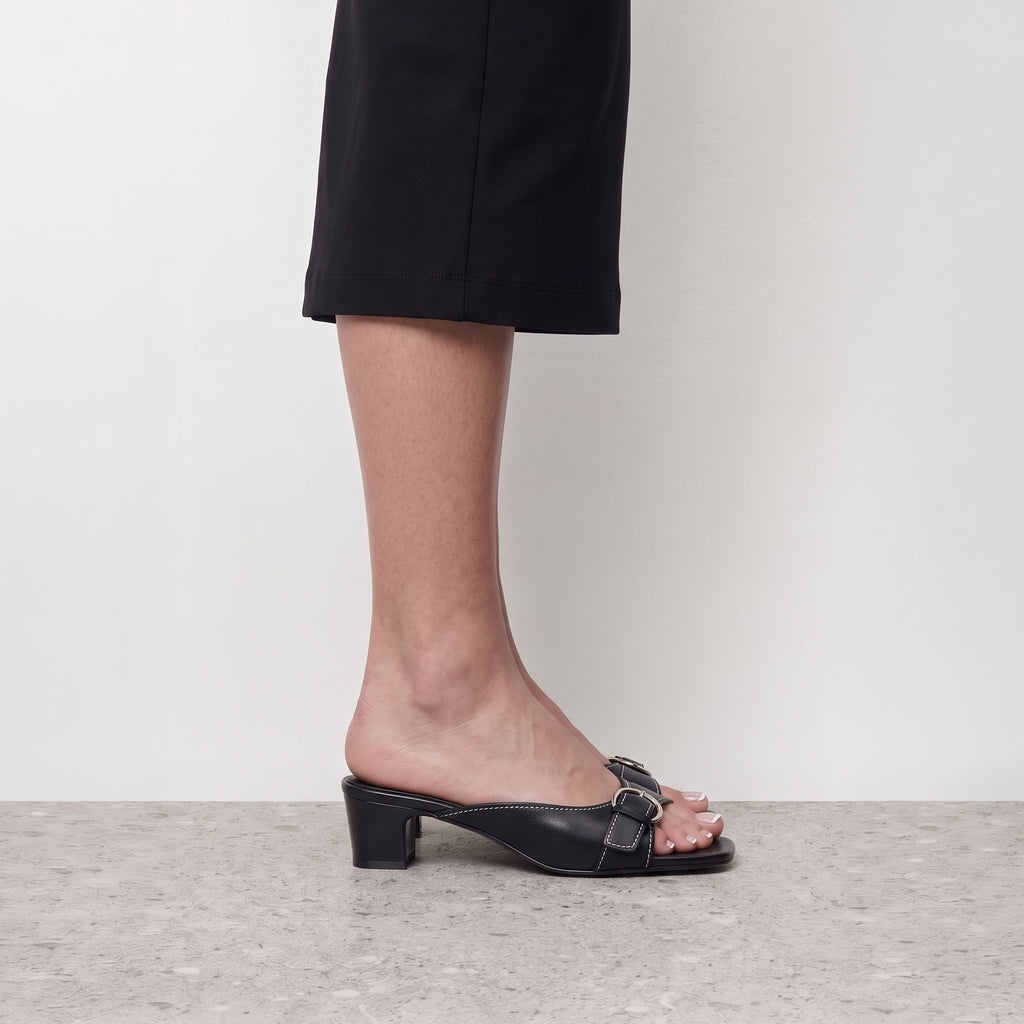 Side profile of a black buckle mule heel worn with a black skirt