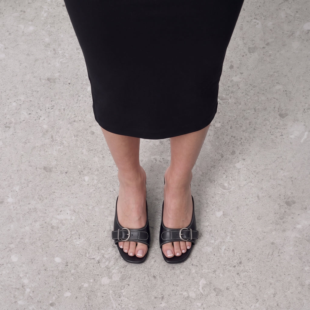 Front view on the foot of black buckle mules on a low block heel