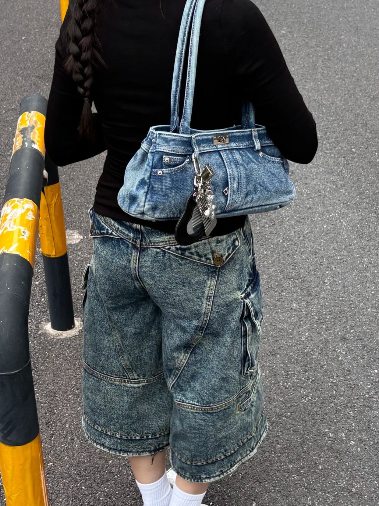 Person wearing a black shirt and blue denim overalls with a blue handbag, standing on a gray pavement.