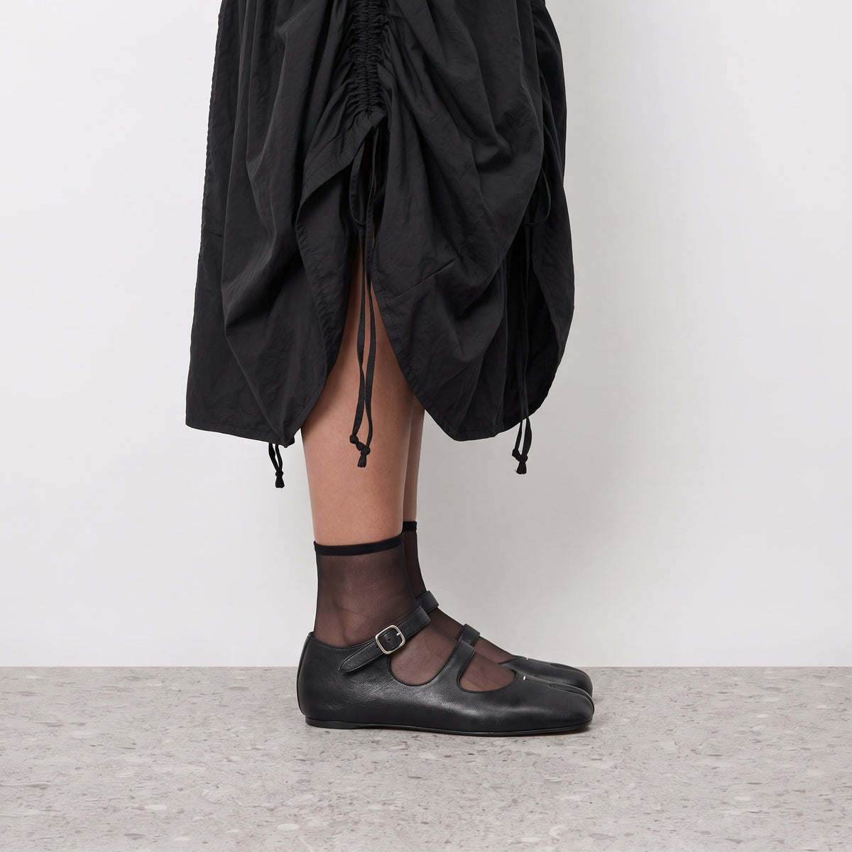 Side profile of a black double strap tabi mary jane worn with sheer socks and a black skirt