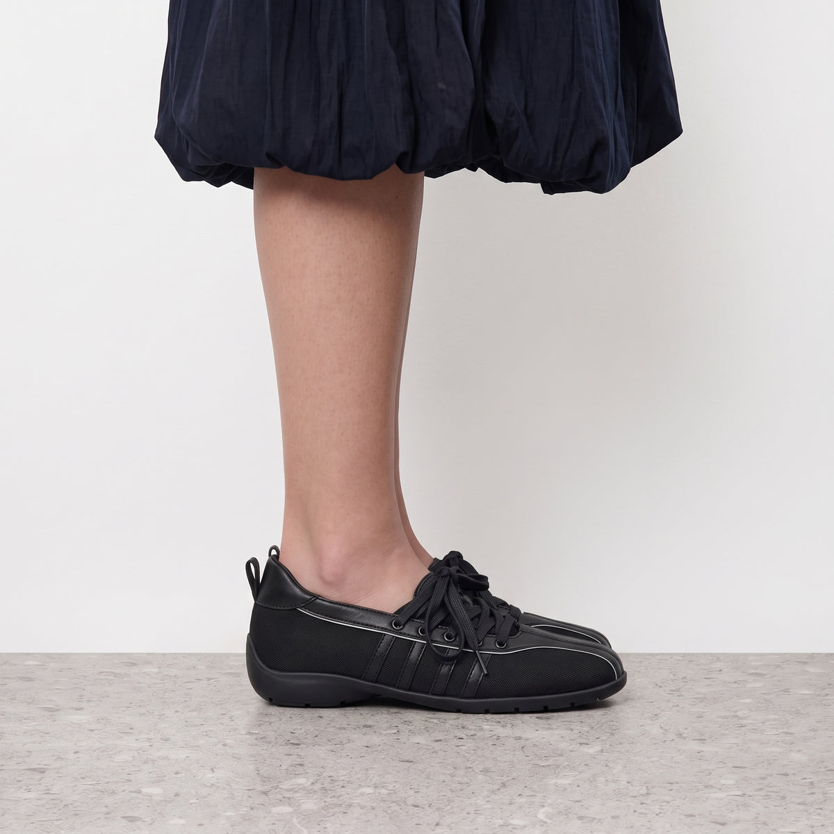 Side profile of a black Iris lace-up sneaker flat worn with a navy bubble-hem skirt