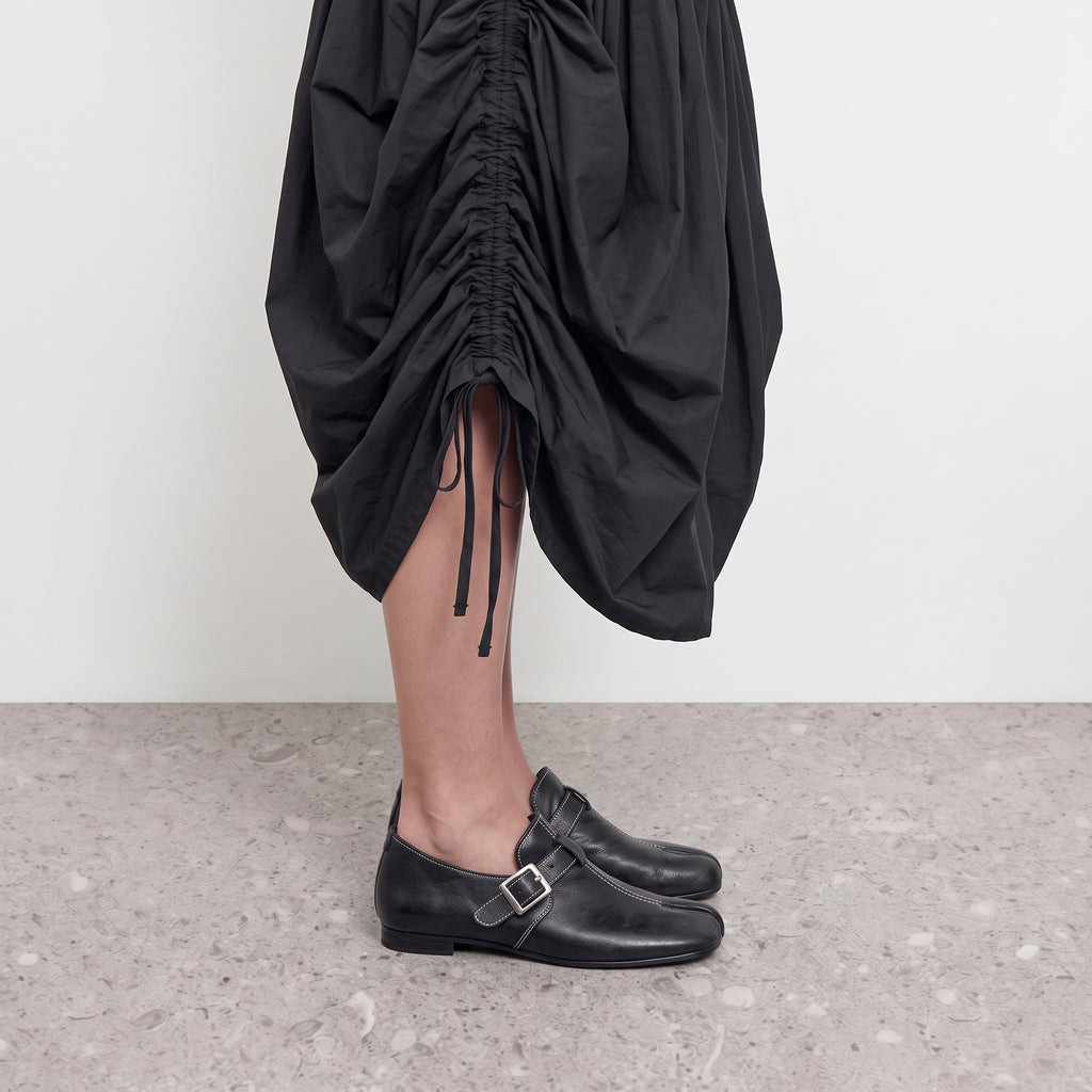 Side profile of a black tabi loafer with a monk strap worn with a ruched black skirt