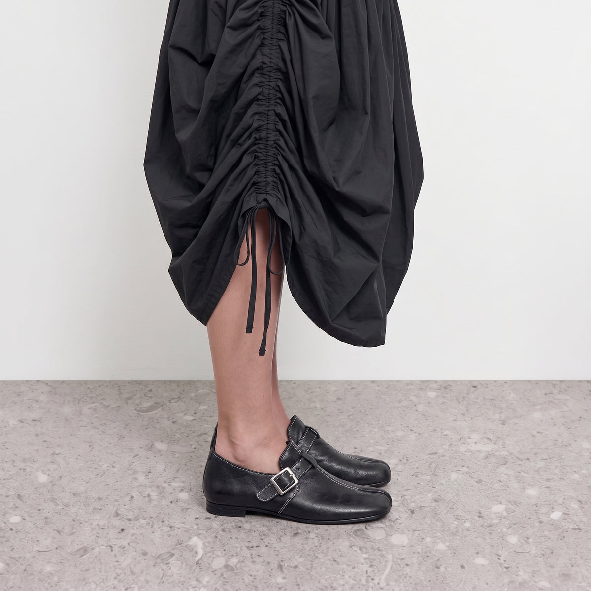 Side profile of a black tabi loafer with a monk strap worn with a ruched black skirt