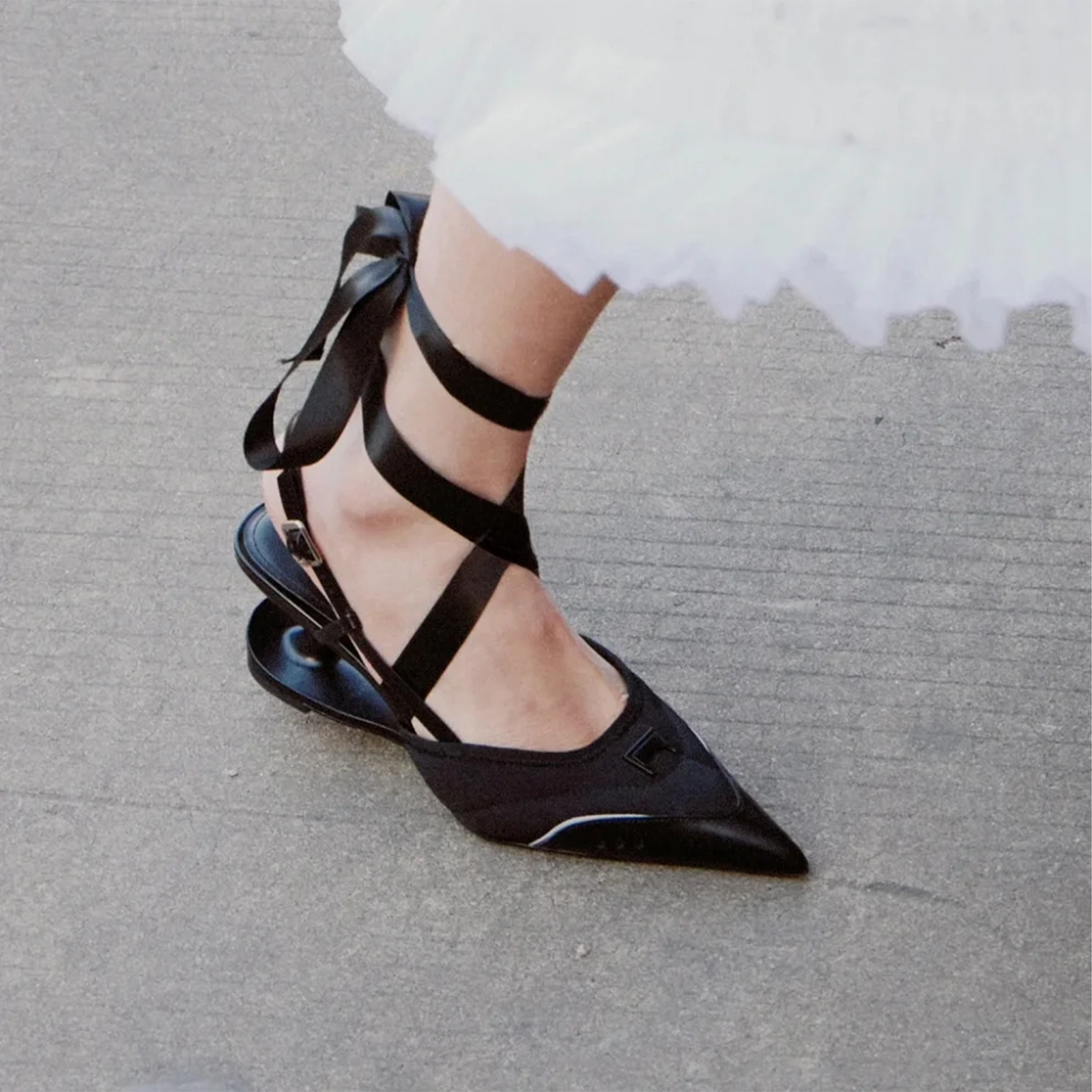 Black pointed-toe Ballerina heels with ribbon straps worn by a person on a gray pavement.