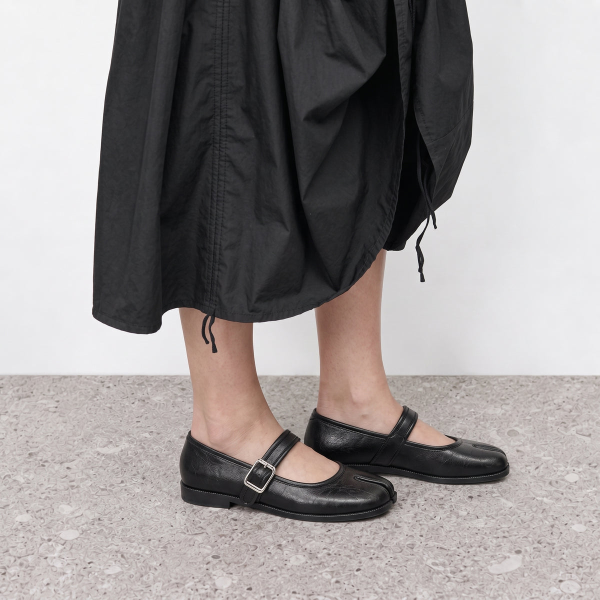Side profile of a black tabi mary jane flat worn with a full black skirt
