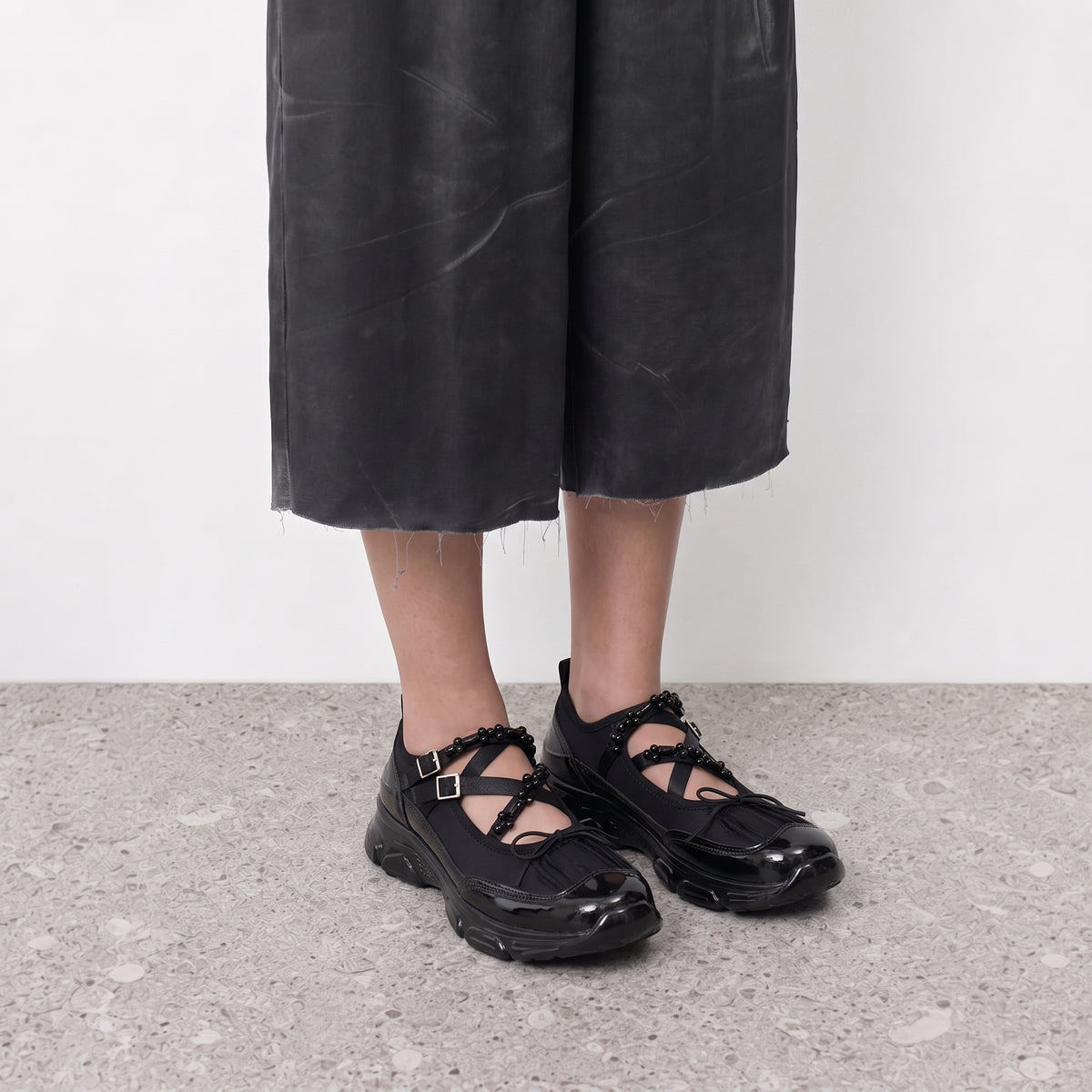 Model wearing black chunky ballet sneakers with crossed bead straps on a stone floor